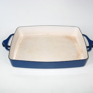 May include: A rectangular baking dish with a white interior and a blue exterior. The dish has two curved handles on either side. The interior shows signs of use. This cast iron baking dish is perfect for roasting and baking.