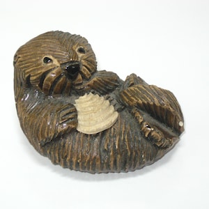 May include: A brown carved wooden figurine of a sea otter lying on its back. The otter has textured fur details, a smooth black nose, and is holding a light-colored, ridged, fan-shaped shell against its chest with both front paws.