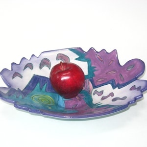 May include: A decorative, leaf-shaped ceramic bowl with a vibrant abstract design in shades of purple, teal, and blue. A red apple rests in the center. The bowl has a scalloped edge.