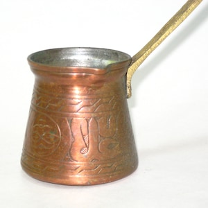 May include: A copper-colored Turkish coffee pot with a brass handle. The pot features etched designs around the body. The handle is attached to the side of the pot. The pot is designed for brewing coffee.