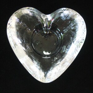 May include: A clear glass heart-shaped dish with a textured surface. The dish has a small, round indentation in the center.