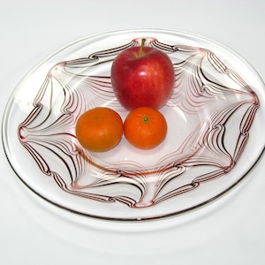 May include: A clear glass serving bowl with a decorative red and black swirl design, holding a red apple and two oranges. The bowl is round with a slightly raised center.
