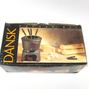 May include: A black Dansk fondue set box featuring an image of a cast iron fondue pot on a stand with long forks, a wedge of cheese, and bread. The packaging displays the brand name Dansk and the text Fondue Set in yellow and white lettering.