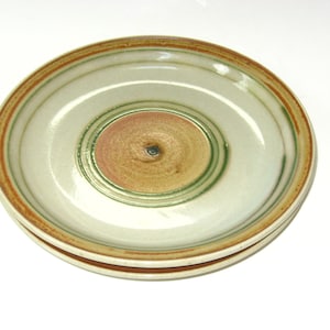 May include: Two ceramic plates stacked on top of each other. The plates have a light beige background with concentric rings of green, brown, and orange. The center of the plates is a darker brown color.