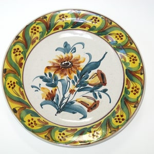 May include: A white ceramic plate with a yellow, green, and brown floral design. The plate has a decorative border with a floral pattern.
