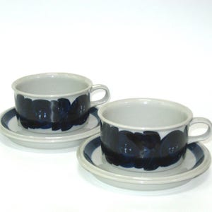 May include: Two ceramic teacups and saucers. The cups and saucers are light gray with a dark blue floral pattern around the cup's rim and saucer's edge. The cups have small handles.