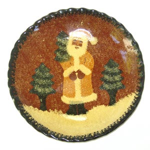 May include: A round ceramic plate with a scalloped edge featuring a painted scene of Santa Claus standing in a snowy landscape between two evergreen trees. The background is a textured reddish-brown, and the base is covered in a sandy beige texture.
