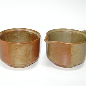 May include: A set of two ceramic serving dishes. One is a round bowl, and the other is a creamer with a handle and spout. Both have a two-tone brown color scheme with a lighter brown on one side and a darker brown on the other.