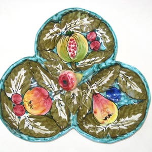 May include: A ceramic serving dish with a three-lobed design, featuring painted fruits and leaves. The dish has a turquoise border and is decorated with a pomegranate, peaches, pears, and berries. The leaves are green and brown.
