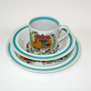 May include: A white ceramic teacup and saucer set with a blue rim and a colorful floral design featuring a yellow house with a red roof.