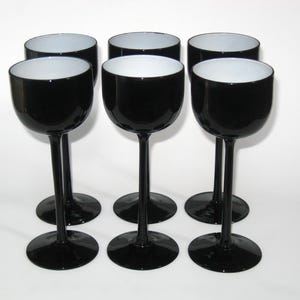 May include: Six black wine glasses with white interiors are arranged on a white surface. The glasses have a rounded bowl shape and a long stem. The base of each glass is circular. The glasses are evenly spaced.