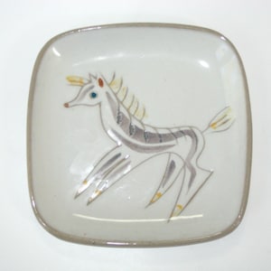 May include: A square ceramic dish with rounded corners, featuring a light gray speckled glaze. The center displays a stylized illustration of a horse in shades of gray, yellow, and blue, with a thin brown border along the outer rim of the dish.