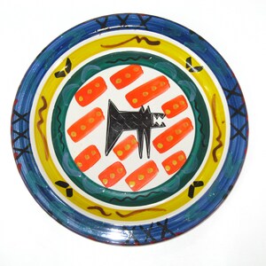 May include: A decorative ceramic plate with a colorful design. The plate features a central white area with a black dog illustration and orange rectangular shapes. The rim has blue, yellow, and green bands with decorative patterns. A unique piece for home decor.