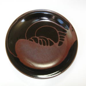May include: A round ceramic plate with a glossy black exterior and a reddish-brown interior. The plate features a dark, abstract design in the center, adding a touch of artistic flair. The plate is suitable for serving food.