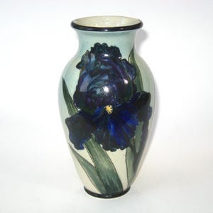 May include: A ceramic vase with a hand-painted iris flower design. The vase features a dark blue iris with green leaves against a light green and cream background. The rim and base are outlined in black.