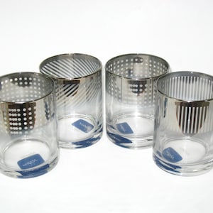 May include: A set of four clear glass tumblers featuring a metallic silver rim and upper band. Each glass displays a distinct pattern, including polka dots, diagonal stripes, and vertical lines, with a blue Mikasa brand sticker on the base.