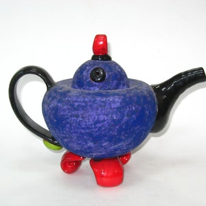 May include: A purple teapot with black handle and spout, red and green accents, and four red legs. The teapot has a textured surface and a black button on the lid.
