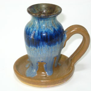 May include: Handmade ceramic vase with a handle and a saucer base. The vase features a gradient of blues and browns, with a dark blue center and lighter blue and brown accents. The handle and base are brown.