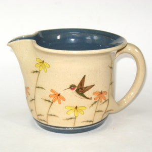 May include: A ceramic pitcher with a blue rim and handle. The pitcher is decorated with a hummingbird and flowers in shades of yellow, orange, and green.