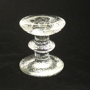 May include: Clear glass candlestick holder with a textured surface. The candlestick has a round base, a central stem with a bulbous section, and a top designed to hold a candle. The glass has a slightly frosted appearance.
