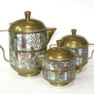 May include: A vintage tea set with a brass finish and iridescent abalone shell inlays. The set includes a teapot, a creamer, and a sugar bowl, all with matching lids and handles. The tea set is a decorative item.