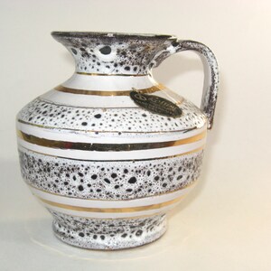 May include: A ceramic pitcher with a handle, featuring a white base with black speckled detailing and gold horizontal stripes. The rim and handle are dark brown. A small tag hangs from the pitcher.