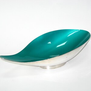 May include: A teal and silver decorative bowl with a leaf-like shape. The interior of the bowl is a glossy teal color, while the exterior is silver. The bowl sits on a small, circular base.