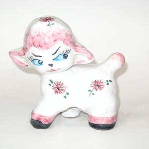 May include: A white ceramic figurine of a lamb with pink painted fur, blue eyes, and green and pink floral details.