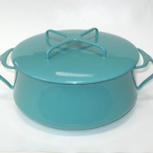 May include: A round, teal-colored enameled metal pot with a matching lid. The lid features a distinctive cross-shaped handle, and the pot has two curved side handles. The exterior surface is smooth, glossy, and uniform in its vibrant teal color.