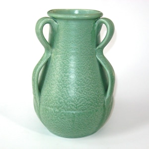 May include: A sage green ceramic vase featuring a bulbous body, a wide circular rim, and two symmetrical curved handles extending from the neck to the mid-section. The surface has a textured, matte finish with a subtle mottled appearance.