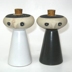 May include: Two ceramic salt and pepper shakers with mushroom-shaped tops and conical bases. The left shaker is white with a black top labeled Salt, and the right is black with a black top labeled Pepper. Both feature cork stoppers and painted eyes.