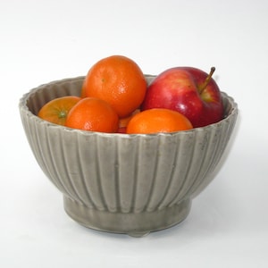 May include: A gray ceramic bowl with a fluted design, filled with oranges and a red apple.