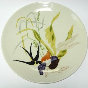 May include: A cream-colored ceramic plate with a hand-painted botanical design. The design features stylized plants in shades of green, yellow, orange, and black, along with purple grapes and a brown fruit. The plate has a glossy finish.