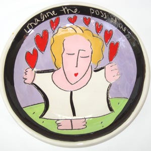 May include: A decorative ceramic plate with a whimsical design. The plate features a stylized figure surrounded by red hearts, with the words "Imagine the possibilities" around the rim. The color palette includes white, black, purple, green, and red.