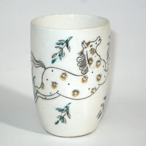 May include: White ceramic tumbler with a hand-drawn design of a running horse. The horse is decorated with yellow floral patterns and black outlines. Green and black leaf accents are also present.