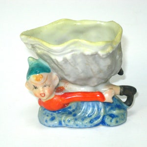 Puede incluir: Una maceta de cerámica vintage con un diseño caprichoso. La maceta tiene la forma de una gran nube con volantes sostenida por un elfo sonriente. El elfo lleva un gorro verde azulado, una camisa roja y pantalones azules. La maceta está pintada en colores pastel.