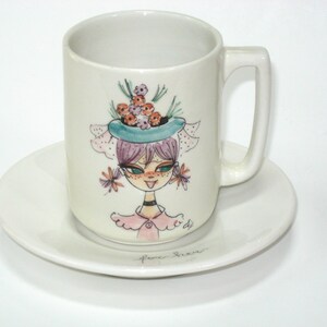 May include: A white ceramic mug and saucer set. The mug features a whimsical illustration of a girl with purple hair, a blue hat, and flowers. The saucer is plain white. The set is perfect for coffee or tea.