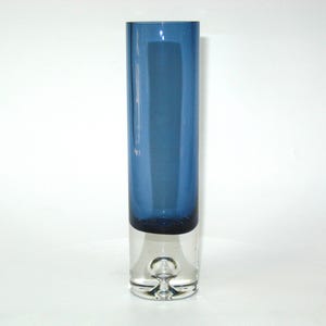 May include: A tall, cylindrical vase with a two-tone design. The upper portion is a deep blue, while the lower section is clear glass. The vase has a rounded base with a small, decorative bubble. The overall design is modern and minimalist.