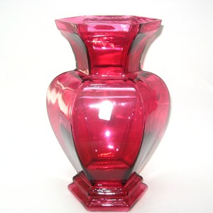 May include: A ruby red glass vase with a geometric design. The vase has a wide, heart-shaped body and a flared, octagonal top. The base is also octagonal. The vase is approximately 10 inches tall.