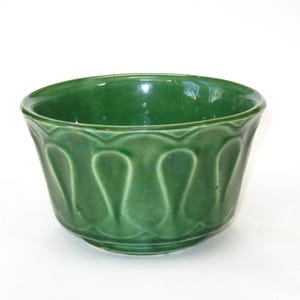 May include: A glossy, emerald green ceramic bowl. The bowl has a scalloped rim and a repeating, stylized leaf pattern around the exterior. The bowl is round with a slightly flared shape and sits on a small base.