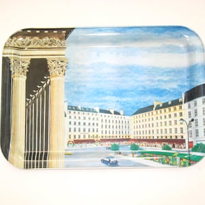 May include: Rectangular metal serving tray with a painted scene of a European city square. The tray features a blue sky, white buildings, and classical columns on the left side. The tray has rounded corners.