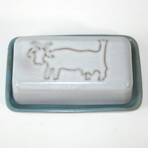 May include: A ceramic butter dish with a light blue base and lid. The lid is white with a brown outline of a cow. The dish is rectangular and has a simple, rustic design.