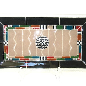 May include: Rectangular decorative glass tray with a black border and a central design featuring a beige background with vertical wavy lines. The center has a black and white circular pattern. The tray is supported by a clear stand.
