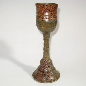 May include: A tall, handcrafted ceramic goblet with a textured, speckled brown rim and a green-brown body. The stem is textured, and the base features detailed relief patterns. The goblet stands approximately 8 inches tall.