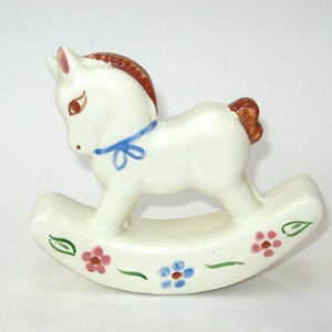 May include: White ceramic rocking horse figurine with brown mane and tail, blue ribbon around neck, and painted pink and blue flowers on the base.