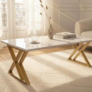 May include: A rectangular coffee table with a white marble top and gold-colored, X-shaped legs. A vase with dried stems, books, and a teacup with saucer are on the table. The table is in a bright, airy living room.