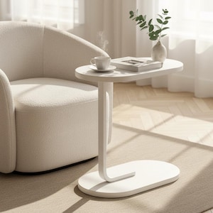 May include: A modern, white side table with an oval top and base, supporting a cup and a book. A white, curved armchair sits nearby. A small vase with greenery adds a touch of nature to the scene. The room has a neutral color palette.