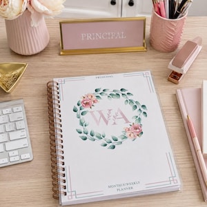 Könnte beinhalten: Ein spiralgebundener Monats- und Wochenplaner mit weißem Einband, verziert mit einem Blumenkranz, den Buchstaben WA und dem Text PRINCIPAL. Der Planer ist umgeben von einer rosa Vase mit Blumen, einem goldenen Namensschild, einem Stiftehalter und einem Hefter.