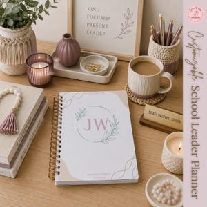 Può includere: Un'agenda a spirale con copertina bianca che mostra le iniziali JW all'interno di una corona di foglie verdi, circondata da accessori da scrivania tra cui una tazza, penne in un portapenne all'uncinetto, una candela, un vassoio e un cartello.