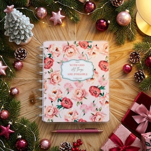 Puede incluir: Una agenda de discos con una cubierta de color rosa claro que presenta un patrón floral de peonías rosas y rojas. La etiqueta central muestra el texto "WITH GOD All Things ARE POSSIBLE". Un bolígrafo rosa a juego yace horizontalmente debajo de la agenda.